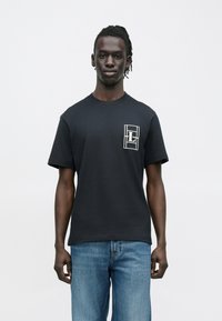 Άνδρας με σκουρόχρωμο δέρμα και πλεξούδες, φορώντας μαύρο μπλουζάκι με λευκό γεωμετρικό λογότυπο "L" στο στήθος και μπλε τζιν, στέκεται μπροστά σε λευκό φόντο.