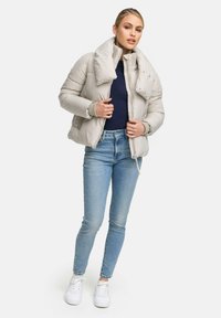 Frau, die eine hellbeige Pufferjacke, einen marineblauen Pullover, hellblaue Jeans und weiße Turnschuhe trägt, vor einem schlichten Hintergrund steht.