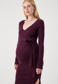 Maternity dress in deep purple ribbed knit, featuring a deep V-neck, long sleeves, and a tied waist belt with thigh-high side slit.