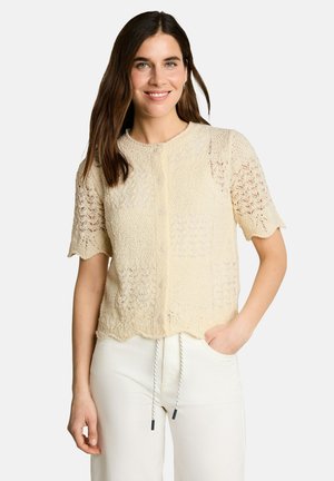 Woman with long brown hair wearing a beige short-sleeve knit cardigan and white pants with drawstring, smiling with one hand in pocket.
