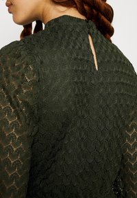 Tmavě zelené krajkové šaty s texturovaným vzorem, vysokým výstřihem a detailem na zapínání na knoflík na zádech se malým otvorem ve tvaru klíče.