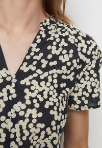 Nahaufnahme einer schwarzen V-Ausschnitt-Bluse mit kurzen Puffärmeln, die ein abstraktes Muster aus weißen Punkten auf dem Oberkörper einer Frau zeigt.