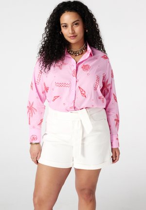 Vrouw met krullend haar die een roze overhemd met rode zeethema-opdrukken en witte short met riem draagt tegen een effen achtergrond.