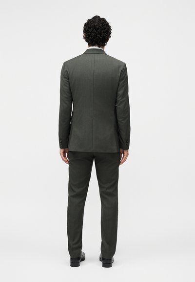 Άνδρας με κατσαρά μαλλιά που στέκεται με την πλάτη προς τα εμπρός, φορά σκούρο γκρι κοστούμι, μαύρα παπούτσια, σε ένα μονόχρωμο ανοιχτό γκρι φόντο.