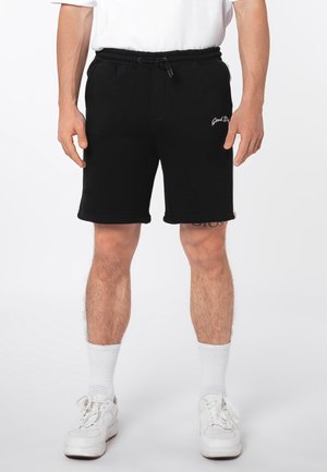 Mann trägt schwarze Shorts mit Kordelzug und dem Schriftzug "Good Style" am linken Oberschenkel, weiße Socken, weiße Sneaker und ein weißes Hemd.