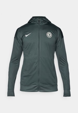 Donkergroene Nike-jack met een volledige ritssluiting aan de voorkant, capuchon, zijzakken en een Chelsea FC-logo op de bovenste linkerborst. Gladde stofstructuur.