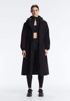 Femeie în picioare purtând o haină lungă neagră cu glugă peste un top sport, colanți și adidași negri, privind înainte pe un fundal simplu.