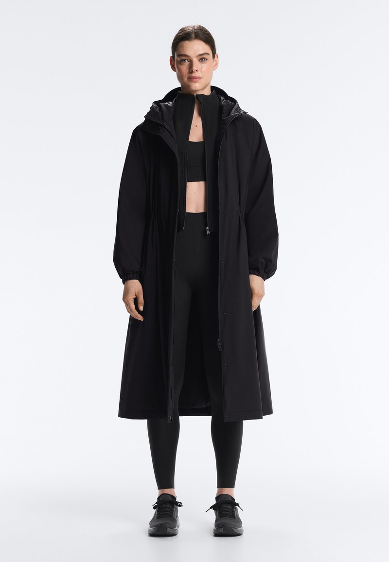 Femme debout portant un long manteau noir à capuche par-dessus un soutien-gorge de sport, des leggings et des baskets noires, face à l'avant sur un fond uni.