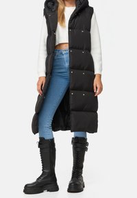 Schwarze Steppweste mit Kapuze, ausgestattet mit einem gesteppten Design, goldenen Druckknöpfen und einer langen Silhouette, kombiniert mit blauen Jeans und schwarzen Kampfstiefeln.