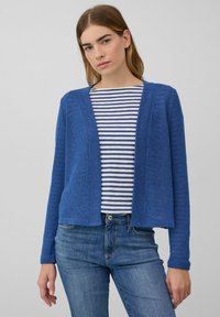Jonge vrouw in een blauwe open cardigan over een wit en blauw gestreept shirt, gecombineerd met blauwe jeans voor een effen achtergrond.