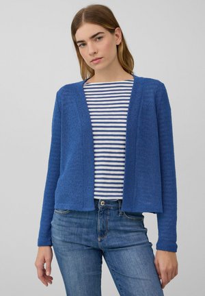 Junge Frau in einem blauen offenen Cardigan über einem weiß-blau gestreiften Shirt, kombiniert mit blauen Jeans, vor einem schlichten Hintergrund.