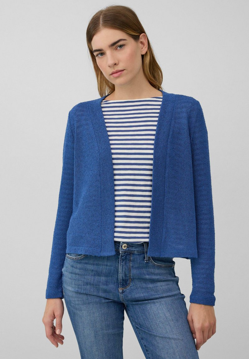 Jonge vrouw in een blauwe open cardigan over een wit en blauw gestreept shirt, gecombineerd met blauwe jeans voor een effen achtergrond.