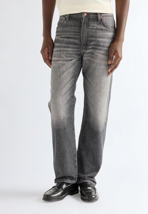 Persona que lleva jeans negros desgastados de pierna recta y mocasines negros de cuero, de pie con las manos relajadas a los lados.