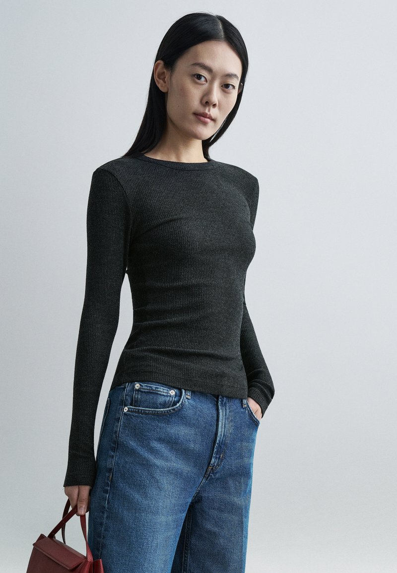 Camisola preta canelada de manga comprida com decote redondo, combinada com jeans de ganga azul. Modelo segura uma pequena mala vermelha.