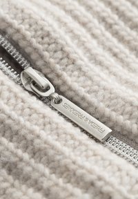 Lichtbeige gebreide stof met een zachte textuur, voorzien van een zilveren metalen rits en een label met de ingekraste merknaam.