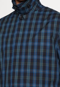 Giacca da uomo con colletto caratterizzata da un motivo a quadri blu navy e verde, con un colletto alto e due bottoni neri visibili. Tessuto liscio.
