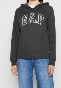 Sweat à capuche gris zippé avec une poche frontale, arborant le logo blanc "GAP" sur la poitrine, capuche avec cordon de serrage et poignets côtelés. Associé à un jean bleu.