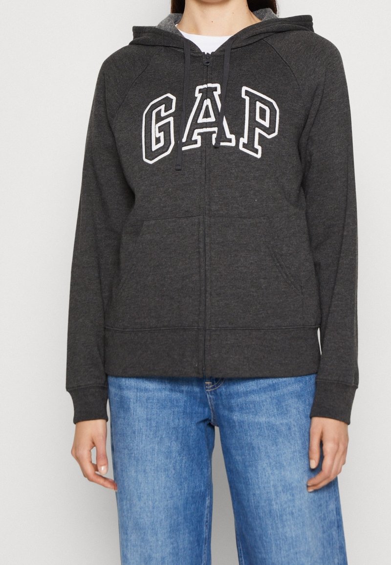 Sweat à capuche gris zippé avec une poche frontale, arborant le logo blanc "GAP" sur la poitrine, capuche avec cordon de serrage et poignets côtelés. Associé à un jean bleu.