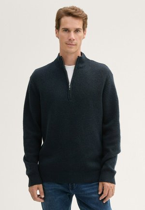 Donker marineblauwe pullovertrui in een zachte, gestructureerde brei met een half-rits kraag en geribde manchetten. Draag deze over een wit T-shirt, gecombineerd met een spijkerbroek.