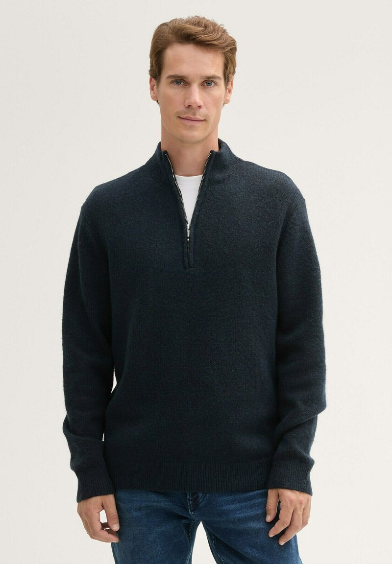 Pull noir marine en tricot doux et texturé avec un col zippé mi-long et des poignets côtelés. Porté sur un T-shirt blanc, associé à un jean.