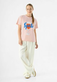 Roze katoenen t-shirt met een blauwe "Spritz" grafiek en glasillustratie, gecombineerd met witte wijde broek en beige sneakers.