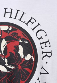 Camiseta de algodón blanca con un diseño circular que presenta patrones florales en rojo y negro, y texto en negrita de color negro que dice "HILFIGER". Textura suave.