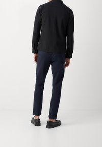 Homme debout, dos tourné vers la caméra, portant une chemise noire à manches longues, un pantalon bleu marine et des chaussures noires sans lacets, sur un fond blanc uni.