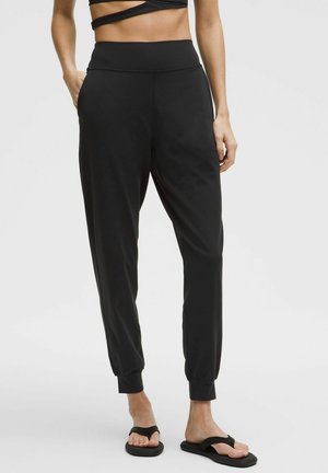 Femme portant un pantalon jogger noir taille haute et des sandales noires, debout avec une main dans la poche devant un fond uni.
