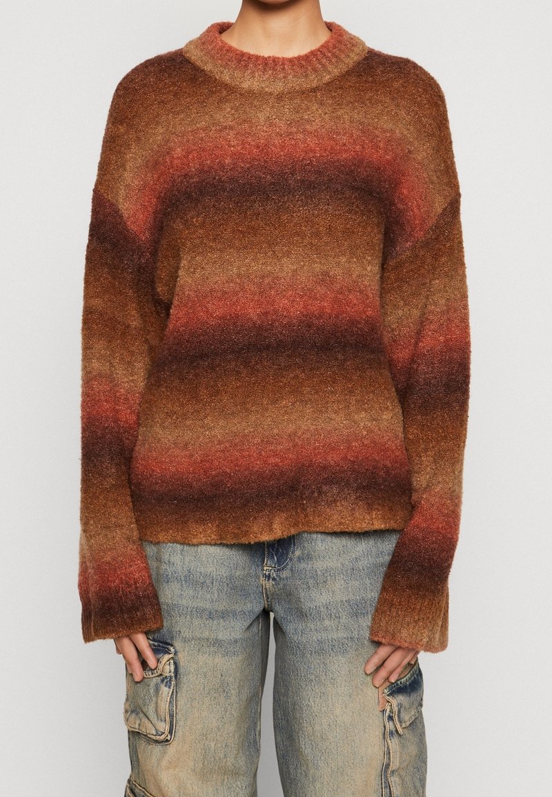 Suéter de punto con cuello redondo, que presenta gradientes horizontales de marrón, óxido y burdeos. Corte holgado y mangas amplias, con una textura suave.