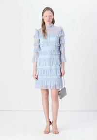 Abito in pizzo azzurro chiaro con strati di volant, maniche lunghe in tessuto trasparente e collo alto. Include una piccola borsa in metallo. Tessuto morbido.