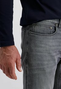 Gros plan sur la main d'une personne à côté d'une poche de jeans en denim gris, portant une chemise à manches longues foncée sur un fond clair.