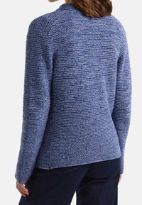 Eine Frau trägt einen strukturierten blauen Strickpullover und dunkle blaue Hose, von hinten vor einem weißen Hintergrund zu sehen.