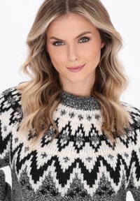 Blonde Frau mit welligem Haar, die einen schwarz-weiß-grauen Strickpullover trägt, der ein geometrisches Muster auf der Brust und den Schultern aufweist.