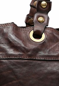 Close-up van een donkerbruine leren tas met gestikte naden, messing ring en band met messing knopen op een witte achtergrond.