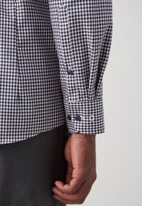 De hand van een man is ontspannen aan de zijkant, gekleed in een zwart-wit geblokt overhemd met lange mouwen, donkere knopen en donkergrijze broek.