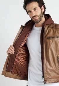 Ruskea nahkatakki, jossa on tilkkutäkkimäinen ruskea vuori, hupulla ja vetoketjusekä. Yksityiskohtainen ompelu olkapäissä ja sivutaskussa.