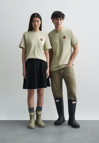 Μαλακά πράσινα βαμβακερά t-shirts με μικρό κομμένο λογότυπο, σκούρα φούστα, μπεζ καμπάνες από βελούδο, πράσινα μποτάκια μέχρι τον αστράγαλο και μαύρες ψηλές καουτσούκ μπότες.