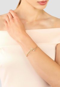 Arany karperec elegáns kialakítással, kis kristály díszítéssel és ívelt formákkal, világos bőrű karon viselve.