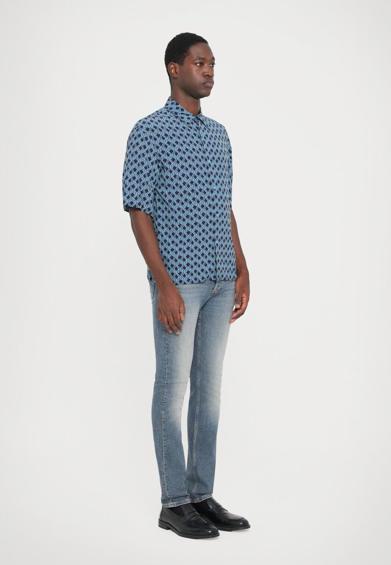 Hombre de pie mirando a la derecha, vestido con una camisa de manga corta azul con estampado, jeans ajustados desgastados y mocasines de cuero negros, sobre un fondo blanco.