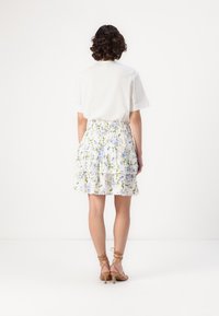 Weiße Bluse mit kurzen Ärmeln und Fransen detaillierte, kombiniert mit einem knielangen, floral gemusterten Rock in weißen, blauen und grünen Farbtönen.