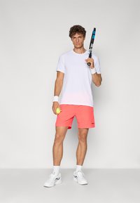 Camisa atlética branca de mangas curtas, calções corais vibrantes com logótipo, segurando uma raquete de ténis e uma bola. Ténis brancos com detalhes pretos.