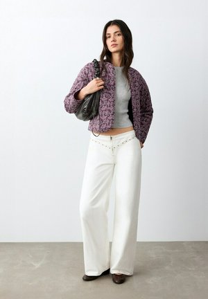 Donna in piedi contro un muro bianco, indossa una giacca viola con motivo, un top grigio corto, pantaloni bianchi larghi con vita borchiata, scarpe marroni, tiene in mano una borsa nera.