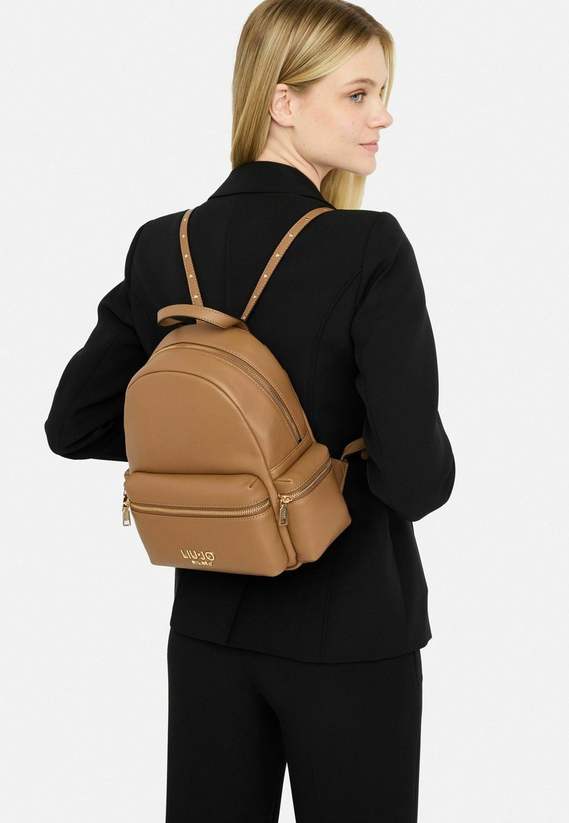 Zaino in pelle marrone con forma arrotondata e due scomparti con zip. Presenta spallacci regolabili con dettagli in borchie e un logo.