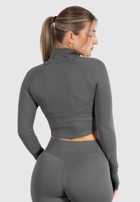 Graues, tailliertes Sportoberteil mit langen Ärmeln, gefertigt aus glattem Material. Es verfügt über einen hohen Kragen und einen figurbetonten Schnitt, kombiniert mit passenden, hochtaillierten Leggings.