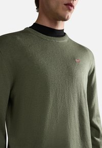 Oliven grüner Strickpullover mit rundem Halsausschnitt und strukturierter Oberfläche. Mit einem kleinen gestickten Flaggenmotiv auf der Brust.