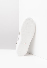 Sneaker bianco con dettagli dorati, suola in gomma testurizzata e design piatto. Presenta il logo e il dettaglio della bandiera italiana sul lato della suola.