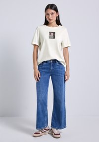 Camiseta de algodón beige clara con un estampado gráfico cuadrado, combinada con pantalones vaqueros de pierna ancha en denim azul y zapatillas multicolores.