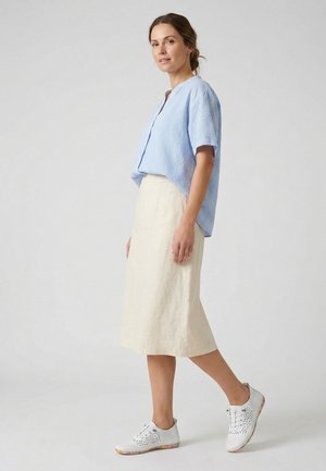 Vrouw staat, draagt een lichtblauw shirt met korte mouwen, een beige midi-rok en witte geperforeerde sneakers met een bloemenpatroon op de zolen.