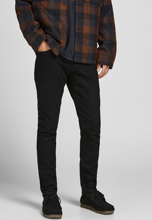 Mężczyzna ubrany w czarne dopasowane dżinsy, czarne buty oraz brązowo-czarną kratowaną koszulę zapinaną na guziki, z rękami w kieszeniach.