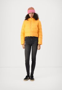 Veste puffer jaune vif courte avec des coutures horizontales, associée à un jean skinny noir et à un bonnet en tricot rayé rose. Baskets noires visibles.